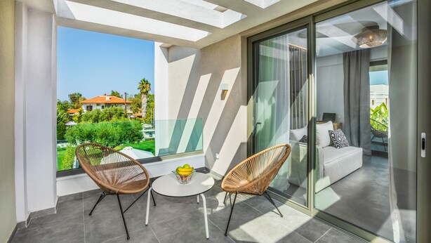 Un balcón con dos sillas de mimbre, una mesita con un frutero, y vista a una zona residencial.