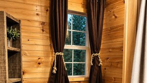 L’intérieur d’un chalet en bois, avec une fenêtre, des rideaux, une étagère avec des serviettes et un petit meuble.