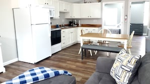 Eine kompakte Küche mit weißen Schränken, ein Essbereich mit Holztisch und -stühlen sowie ein graues Sofa mit gemusterten Kissen.