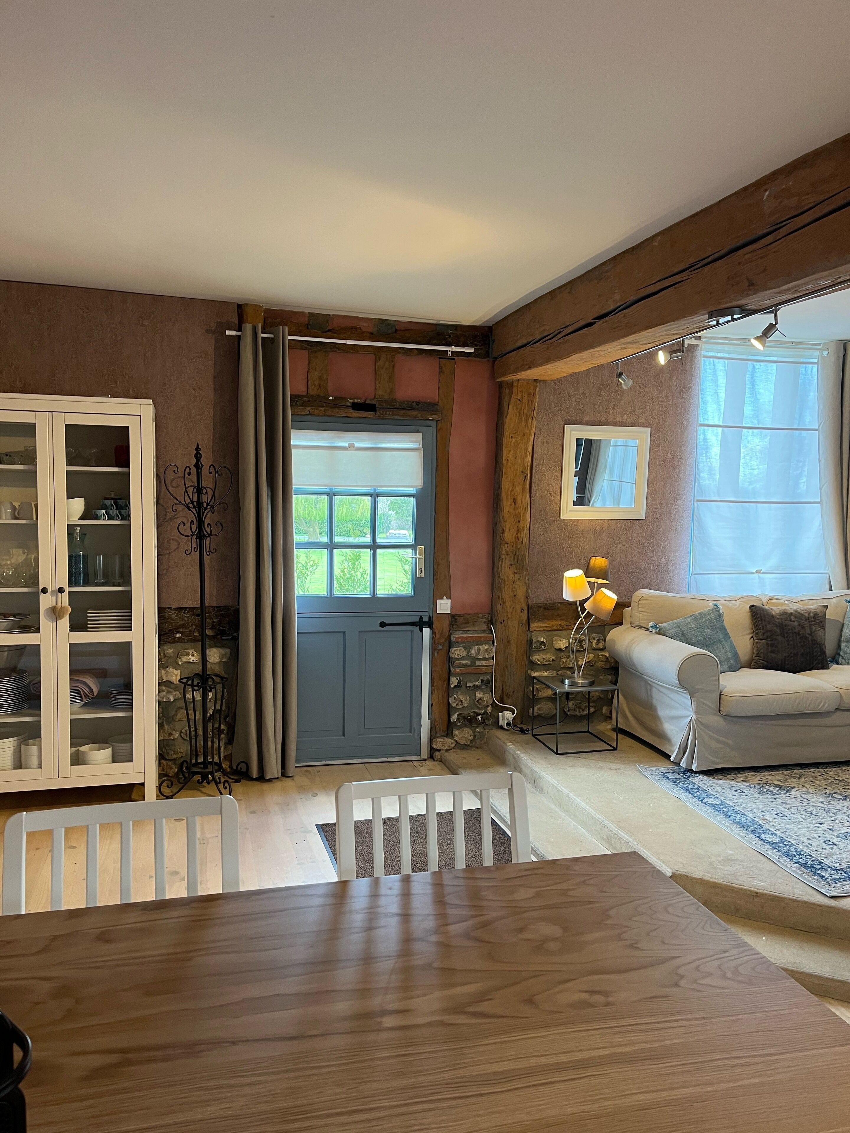Un salon chaleureux avec une table à manger en bois, un buffet blanc aux portes vitrées, une porte bleue et un mur en pierre.