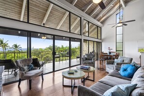 A spacious living area with a large glass balcony, modern furniture, and a view of lush greenery. - Summer Deal Kona Golf View Vacation Rental with Pool and Ocean Views (Kailua-Kona)