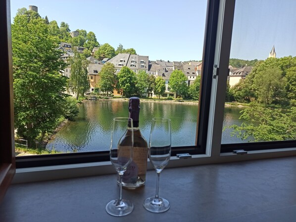 A bottle of wine and two glasses on a table by a window overlooking a river and buildings.
