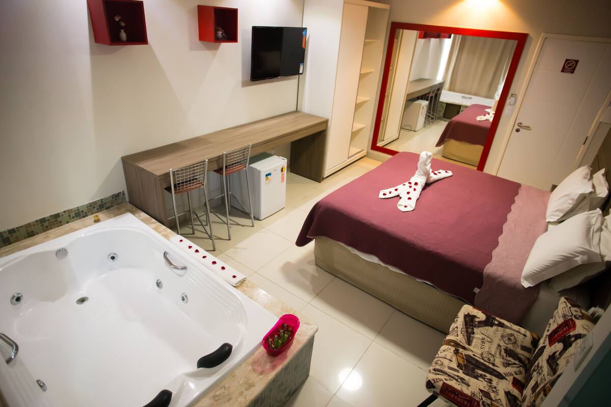 A hotel room with a large bathtub, a bed with a red blanket, a TV mounted on the wall, and a wardrobe with a mirror.