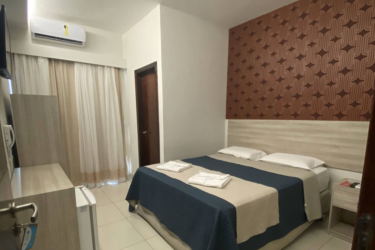 A hotel room with a bed, a desk, an air conditioning unit, and a patterned wall.