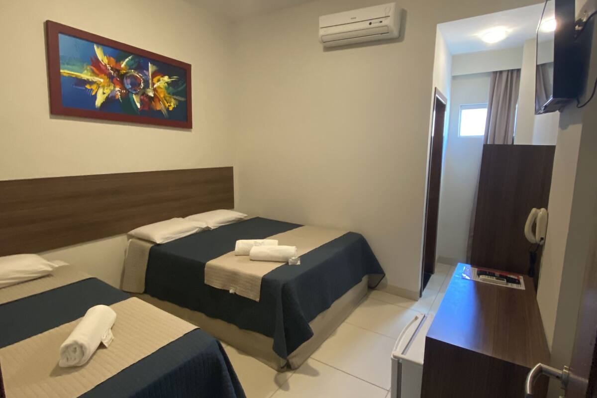 A hotel room with two beds, a desk, a TV, an air conditioning unit, and a painting on the wall.