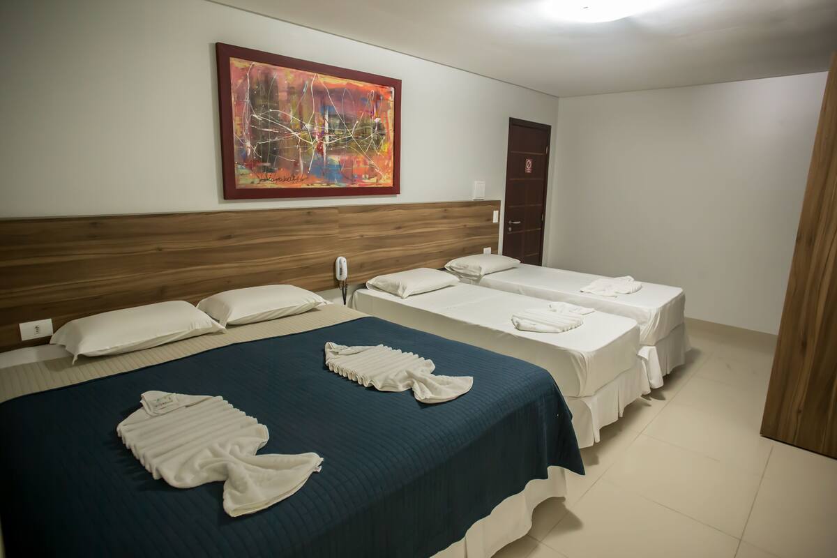 A hotel room with a large bed, two smaller beds, a wooden headboard, a colorful abstract painting, and a door.