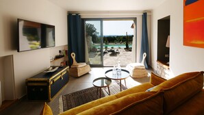 A living room with a sofa, a small table, a television, a suitcase, and a view of an outdoor pool area. - Stone house, private pool, panoramic view, close to the Gorges de l'Ardèche (Orgnac-l'Aven)