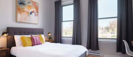 Une chambre moderne avec un grand lit, des rideaux foncés, un luminaire suspendu et une vue sur la ville.