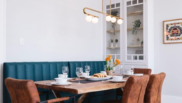 Zona pranzo con tavolo in legno, sedie imbottite marroni, una panca imbottita color petrolio e una lampada a sospensione con tre lampadine.