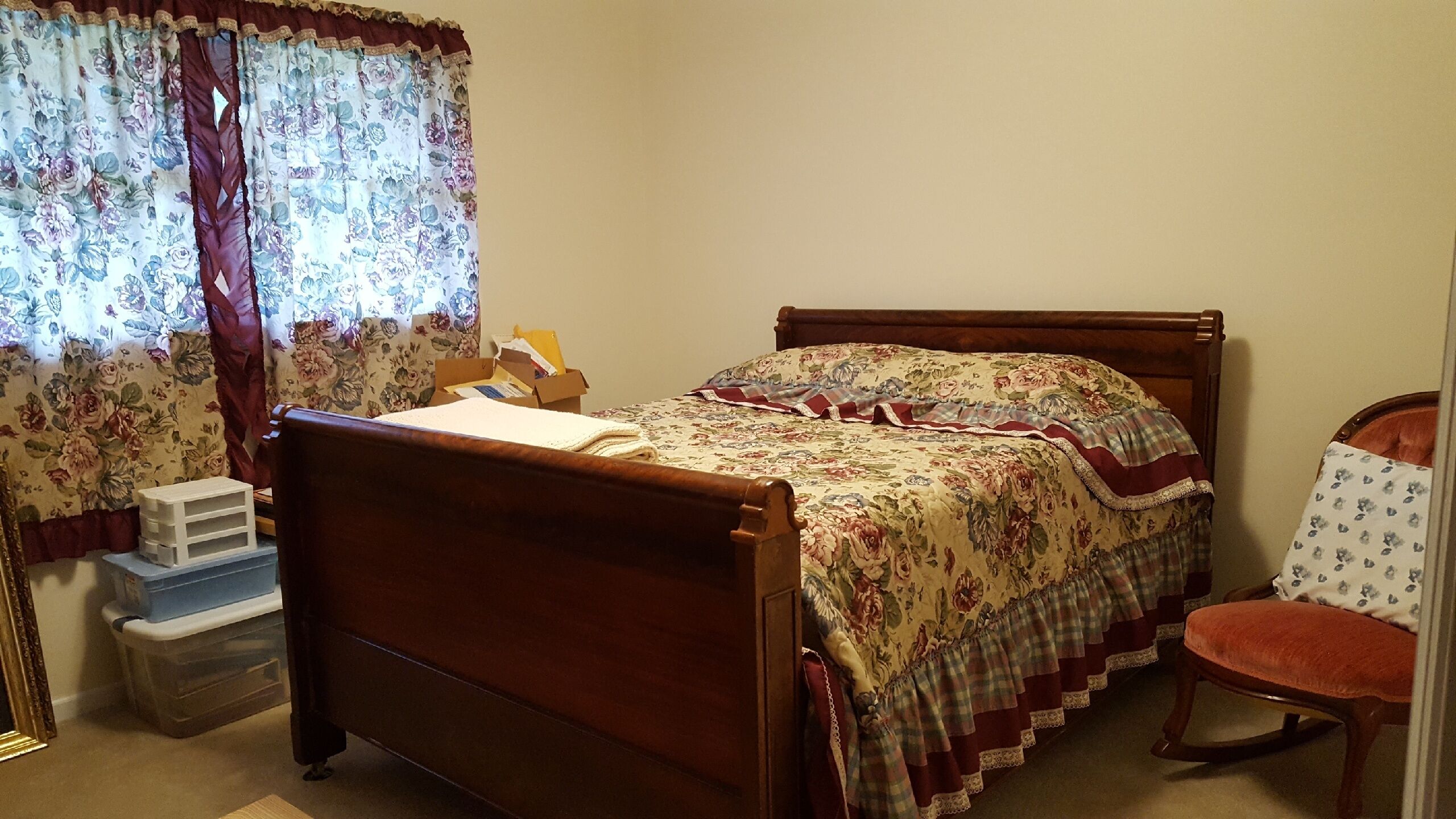 A bedroom with a wooden bed, floral curtains, a wooden chair, and a small table with a box.