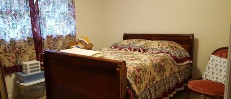 A bedroom with a wooden bed, floral curtains, a wooden chair, and a small table with a box.