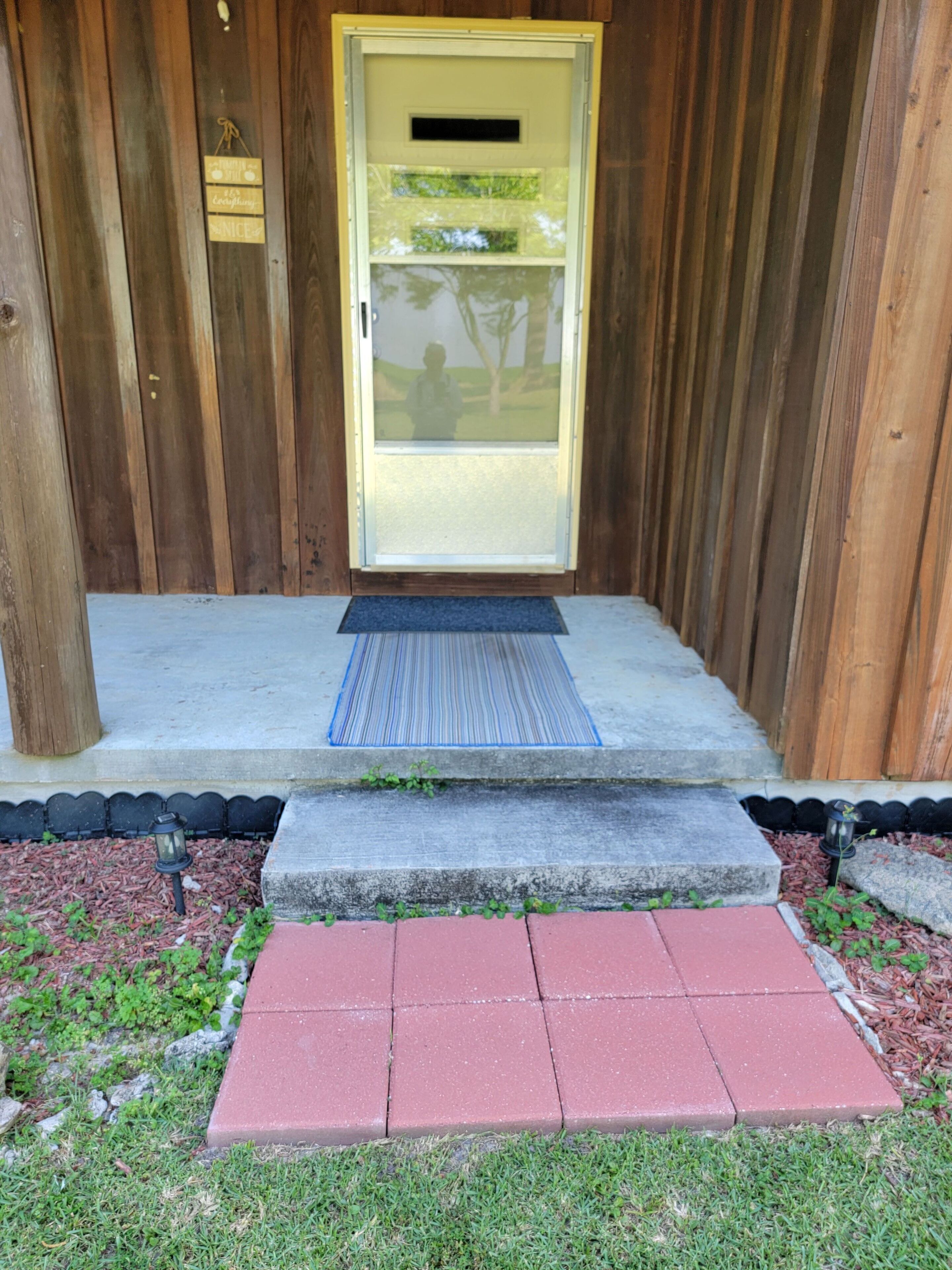 Una puerta de madera con un panel de vidrio, un felpudo y un escalón pequeño que lleva a un porche de concreto.