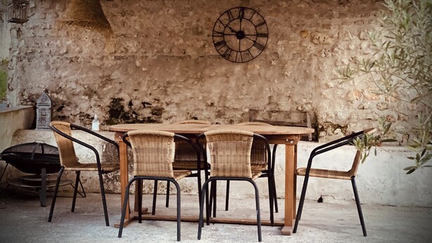 Un espacio comedor al aire libre de estilo rústico, con pared de piedra, mesa de madera y sillas de mimbre.