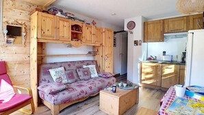 Una sala de estar acogedora con un sofá estampado, armarios de madera, una mesita y una cocina con armarios de madera y un refrigerador.