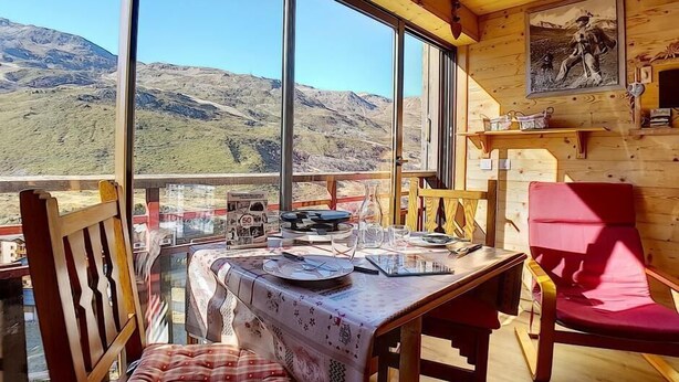 Una cabaña de madera con una mesa de comedor preparada para dos personas, una silla roja y vistas a montañas a través de amplios ventanales.