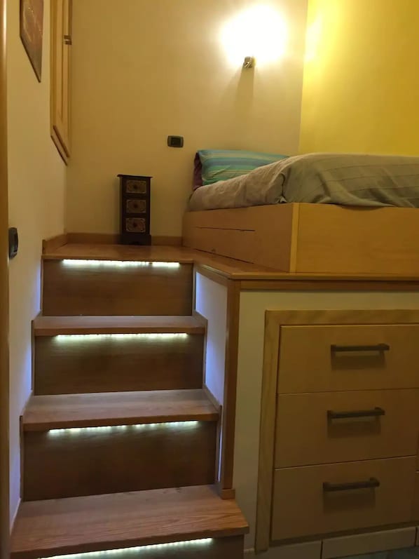 A bedroom with a bed, a nightstand, a dresser, and a staircase with illuminated steps.