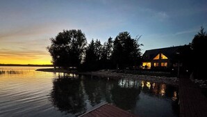 Une propriété au bord du lac, avec un quai en bois, une maison dont l’intérieur est éclairé et des arbres qui se reflètent dans l’eau.