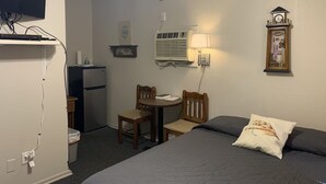 Une chambre d’hôtel compacte comprenant un lit, un petit bureau avec une chaise, un réfrigérateur, un climatiseur fixé au mur, une horloge et une horloge murale.