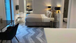 Un dormitorio moderno con una cama grande, un escritorio y una silla. Se ve un espejo, un televisor y un baño al fondo.