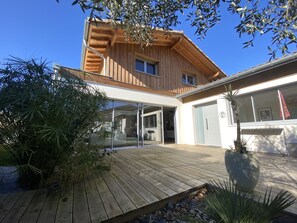 Een modern houten huis met een grote glazen voordeur, een houten terras en veel groen eromheen.