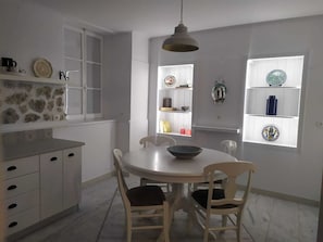 A dining area with a white table and chairs, a hanging light fixture, and shelves with decorative items.