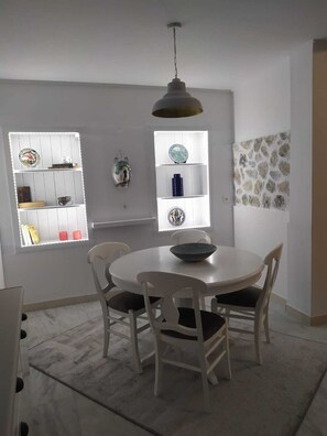 A dining area with a round table and four chairs, a white shelf with decorative items, and a window with a view of the outside.