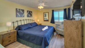 Quarto com cama, mesa de cabeceira, escrivaninha, cadeira, ventilador de teto e janela com cortinas.