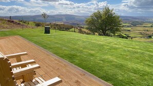 Una terraza de madera con vistas a un campo cubierto de hierba, un árbol y colinas lejanas.