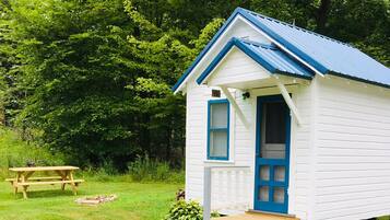 A small white cabin with a blue door and window trim, a wooden bench, and a small porch.