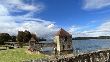 A stone structure with a red roof by a body of water, surrounded by grass and trees.