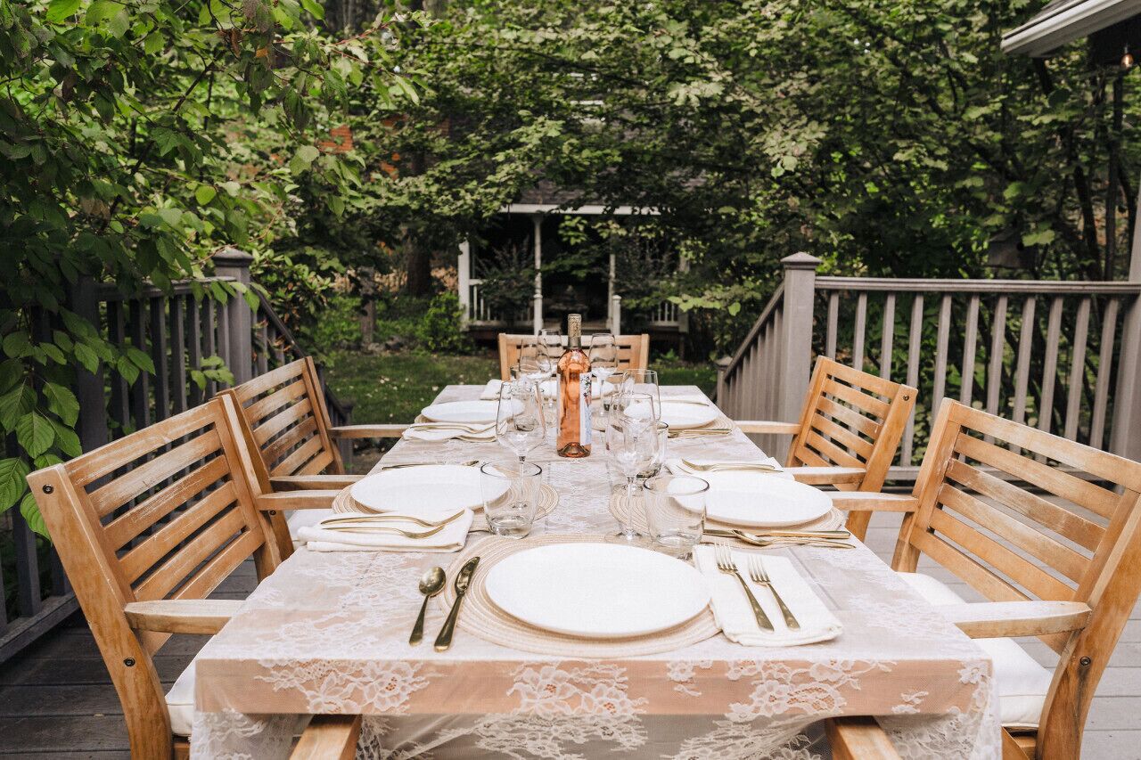 A table set for six with white plates, silverware, and glasses, surrounded by wooden chairs, set on a patio with a wooden railing and greenery in the background.