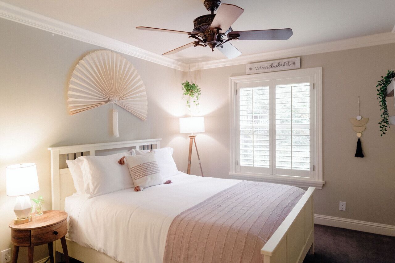 A bedroom with a bed, a bedside table, a lamp, a ceiling fan, a window with blinds, and a wall-mounted sign.