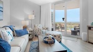 A modern living room with a sofa, ottoman, and chairs, a coffee table, and a view of a balcony with outdoor furniture.