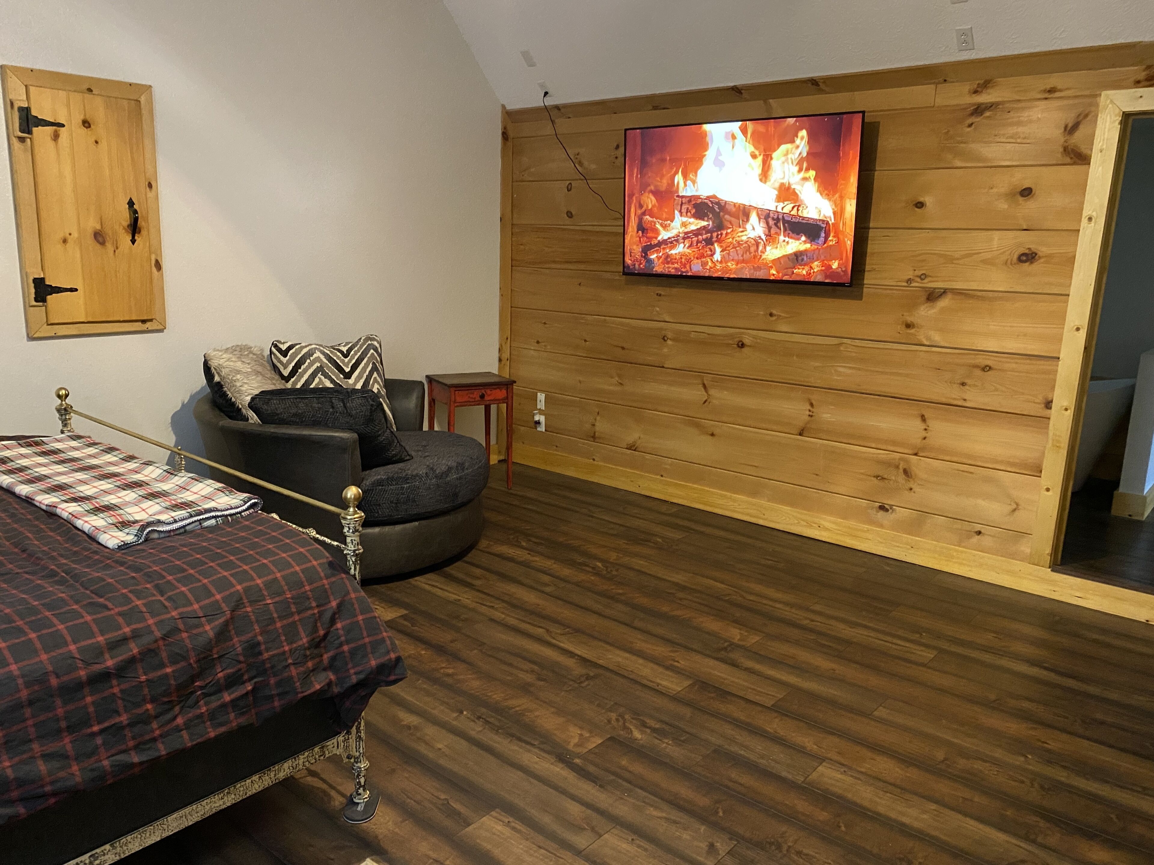 Une chambre confortable avec un lit, un fauteuil, un téléviseur diffusant une image de feu de cheminée et un lambris en bois sur le mur.