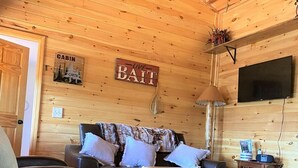 L’intérieur d’une cabane est doté d’un plafond en bois, d’un canapé avec des coussins, d’une table basse, d’un téléviseur et d’une pancarte sur laquelle on peut lire : « APPÂTS VIVANTS ».