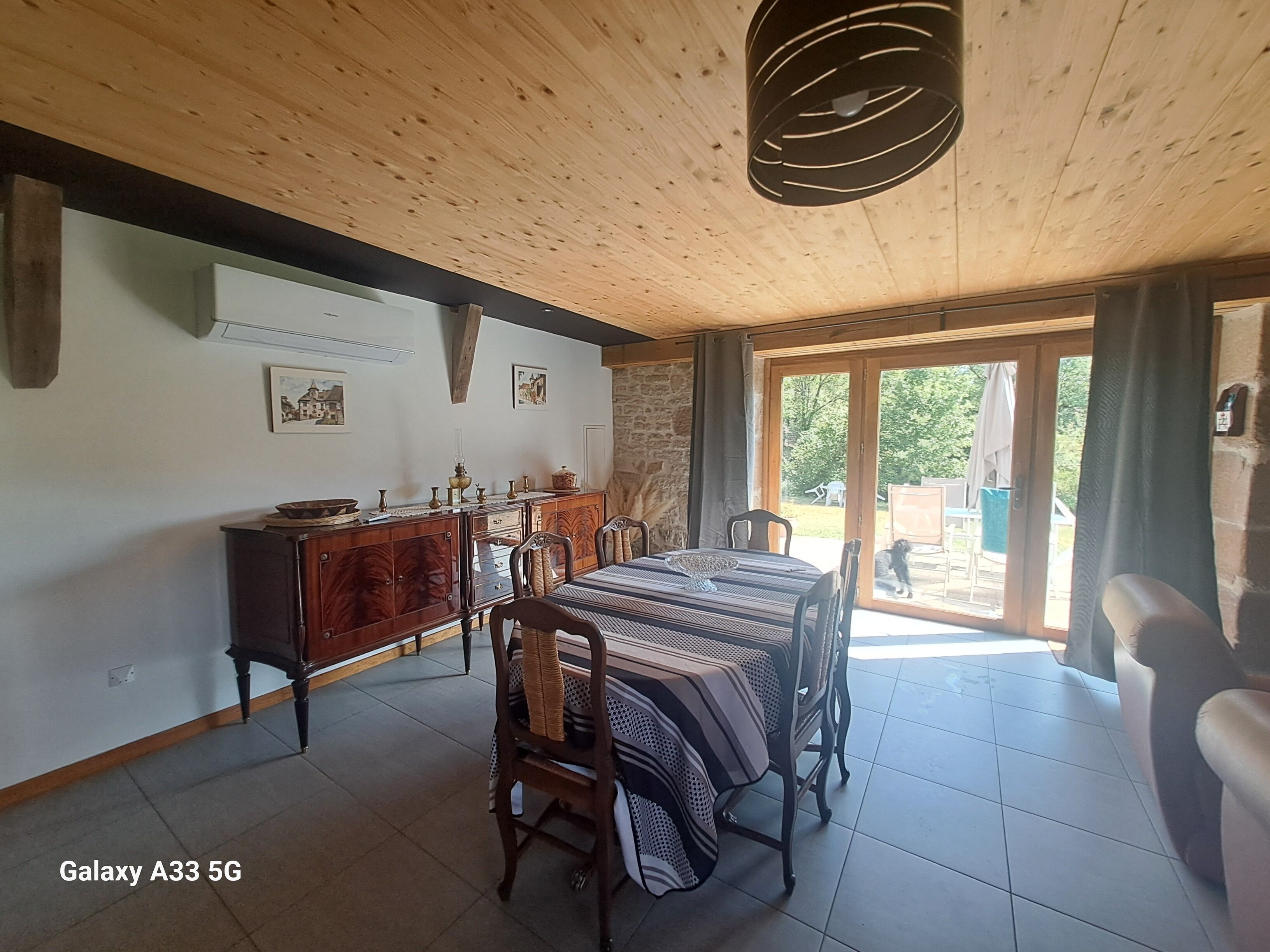 A dining area with a wooden ceiling, a table with chairs, and a view through a glass door to the outside.