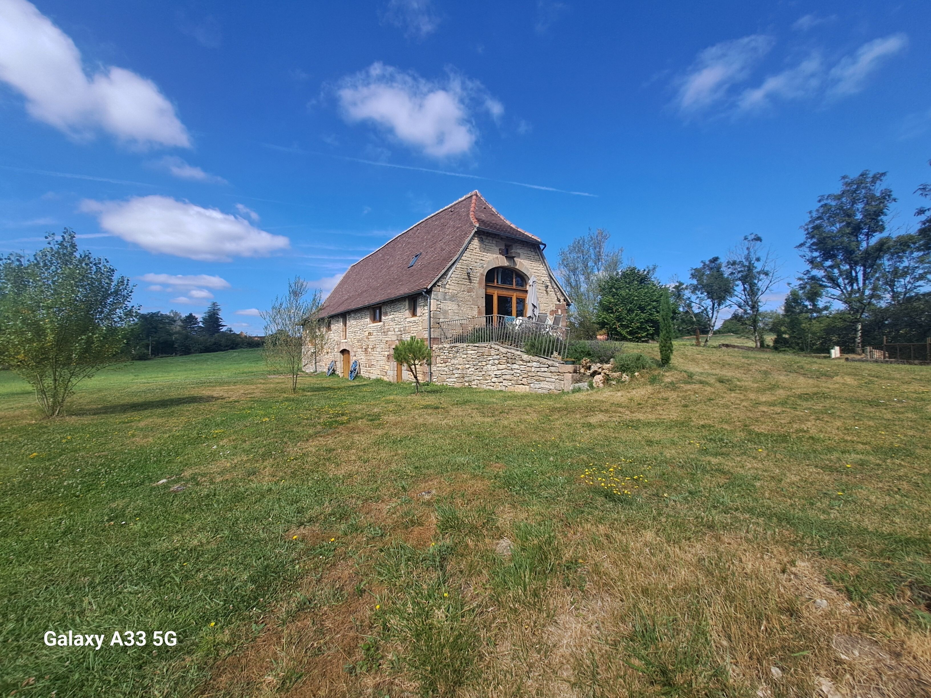 Une maison en pierre avec un toit de tuiles, entourée d’un champ herbeux et d’arbres.