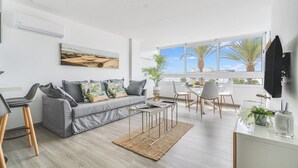 Un salón moderno con un sofá gris, una mesa de centro, un comedor con una mesa y sillas blancas, y un amplio ventanal con vistas a palmeras.