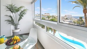 Un balcón con vista a una piscina y edificios, una mesa de comedor con frutas y una planta.