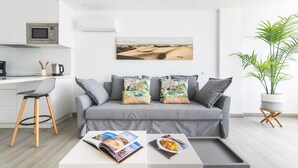 Una sala de estar moderna con un sofá gris, una mesa de centro con revistas, una zona de cocina con microondas y una planta grande.