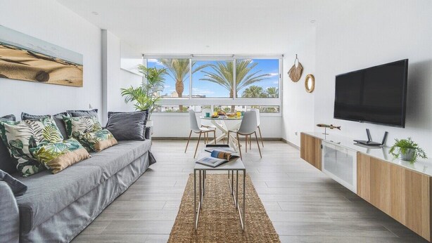 Una sala de estar moderna con un sofá gris, una mesa de comedor y sillas. Un televisor está montado en la pared, y hay una ventana con vistas a palmeras.