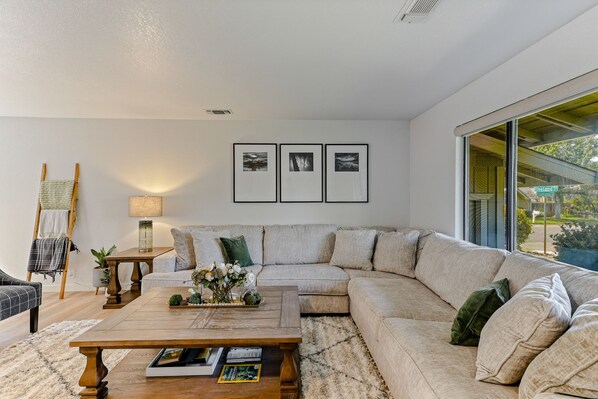 A modern living room with a sectional sofa, a wooden coffee table, and a lamp. There are three framed pictures on the wall and a window with a view of the outside. - ***Pineland Place*** (Redding)