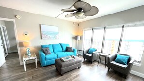 Un salon moderne avec un canapé bleu, un fauteuil gris et un pouf, un ventilateur de plafond et une vue sur le paysage urbain à travers de grandes fenêtres.