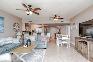 A spacious living room with a sofa, coffee table, and ceiling fans. A kitchen is visible in the background.