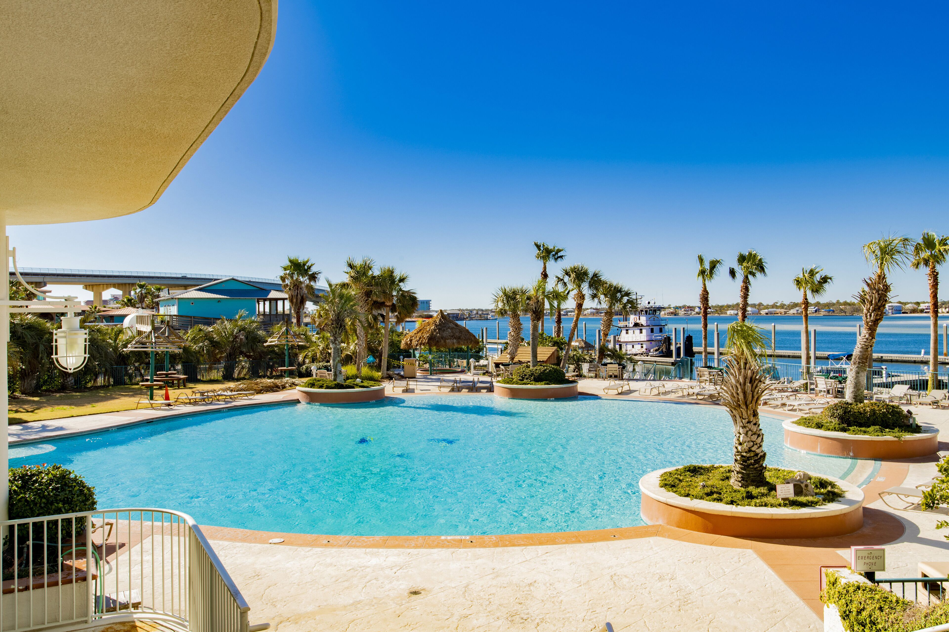 A clear blue pool with palm trees and lounge chairs, surrounded by buildings and a view of the sea.
