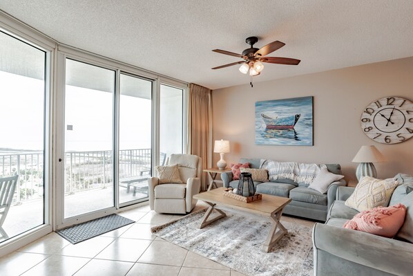 A spacious living room with a balcony, a ceiling fan, a painting of a boat, and a round wall clock. - Beautiful Beach Condo; STUNNING VIEWS OF Gulf & BAY!  (Orange Beach)