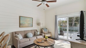 A living room with a sofa, a round coffee table, a ceiling fan, and a large window. - Village Centric Condo w/ Pool Unit 2106 First Floor (St. Simons Island)