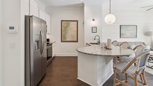 A modern kitchen with a central island, stainless steel appliances, and a dining area with wicker chairs. - Village Centric Condo w/ Pool Unit 2106 First Floor (St. Simons Island)
