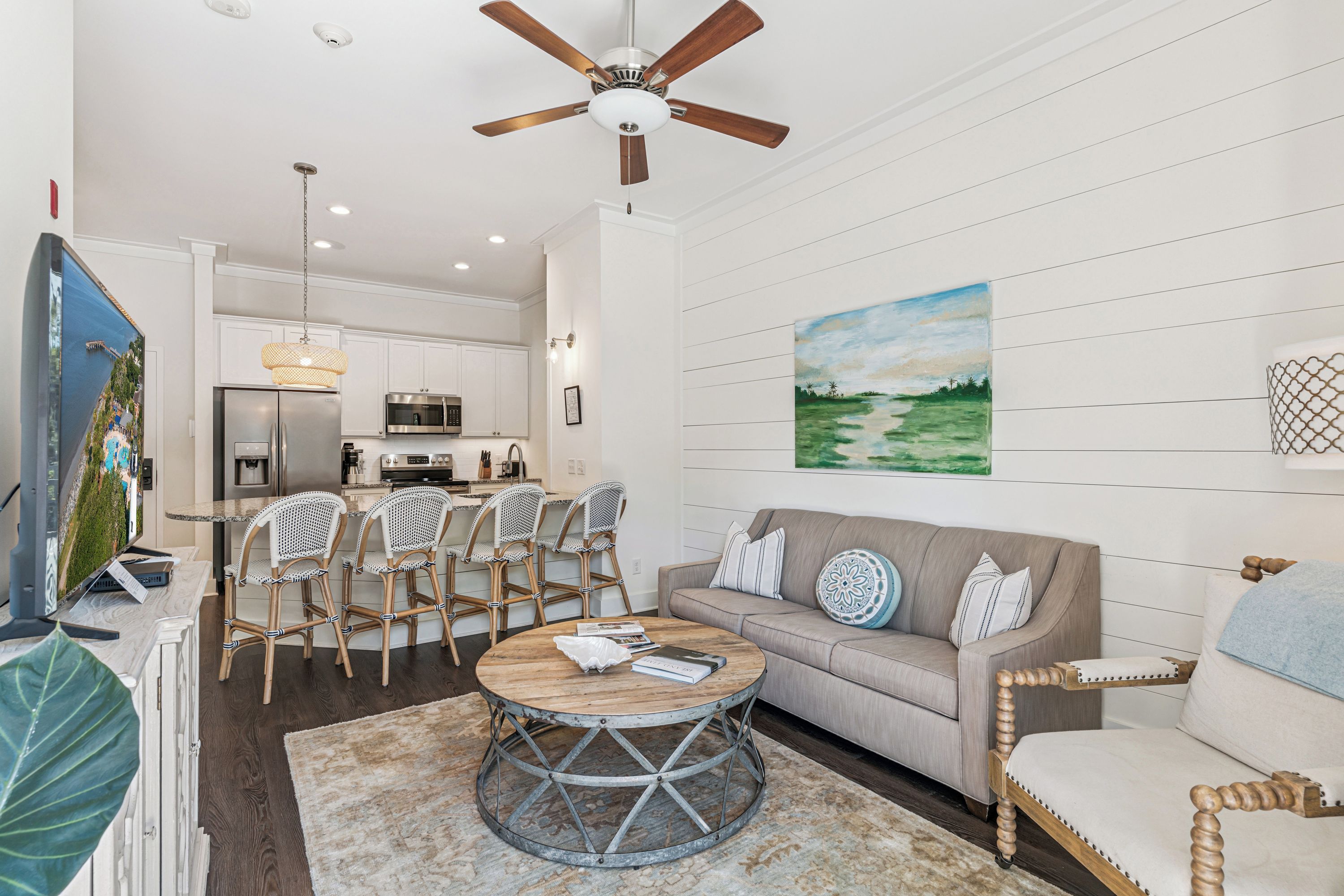 A modern kitchen with white cabinetry, a dining area with wicker chairs, a round coffee table, and a grey sofa with decorative pillows.