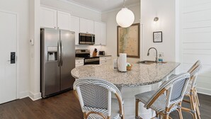A modern kitchen with white cabinetry, stainless steel appliances, a granite countertop island, wicker chairs, and pendant lighting. - Village Centric Condo w/ Pool Unit 2106 First Floor (St. Simons Island)